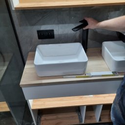 K&K BROTHERS RENOVATION - Montaż czarnej baterii umywalkowej na blacie z drewnopodobnym dekorem, obok biała umywalka nablatowa i złożona miarka stolarska, szare płytki na ścianie.