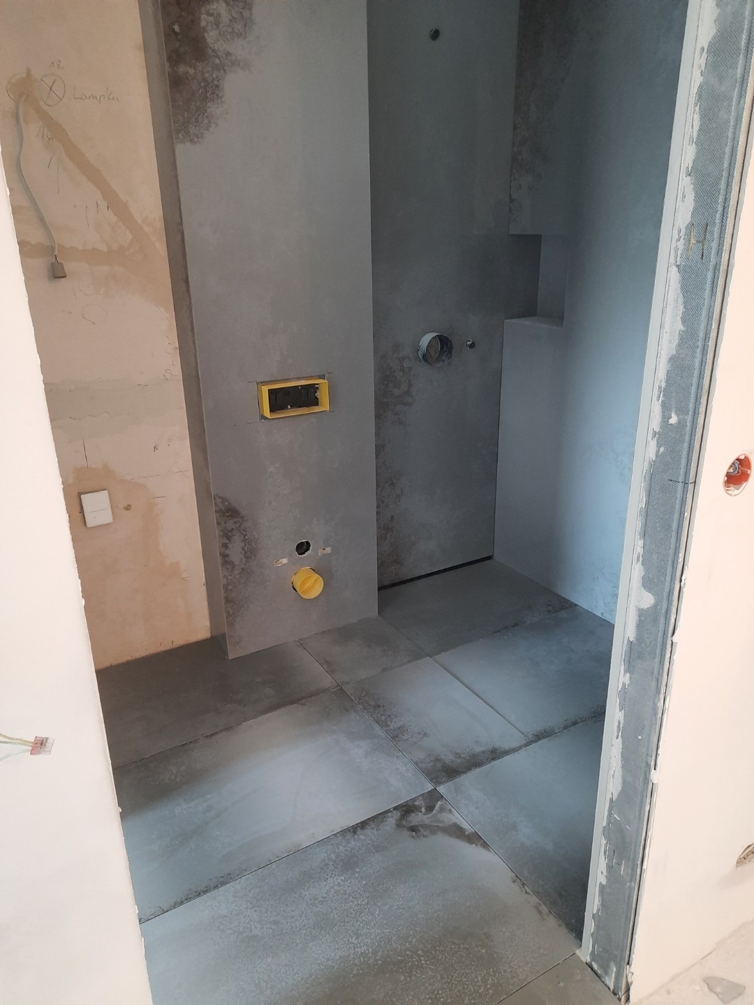 Wnętrze łazienki w trakcie prac wykończeniowych: ułożone szare płytki podłogowe, surowe ściany z widocznymi instalacjami, żółty element instalacyjny i otwór na odpływ, widoczne kable elektryczne...