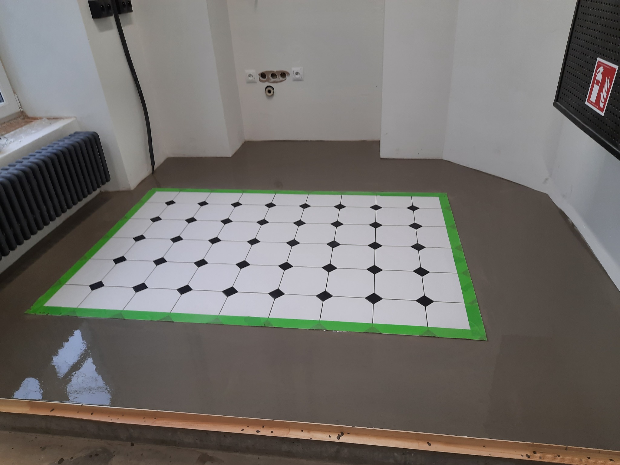 Przygotowanie posadzki w łazience: wylewka samopoziomująca z wyznaczonym obszarem pod płytki w biało-czarną szachownicę, obklejonym zieloną taśmą malarską, widoczne gniazdka elektryczne i grzejnik...
