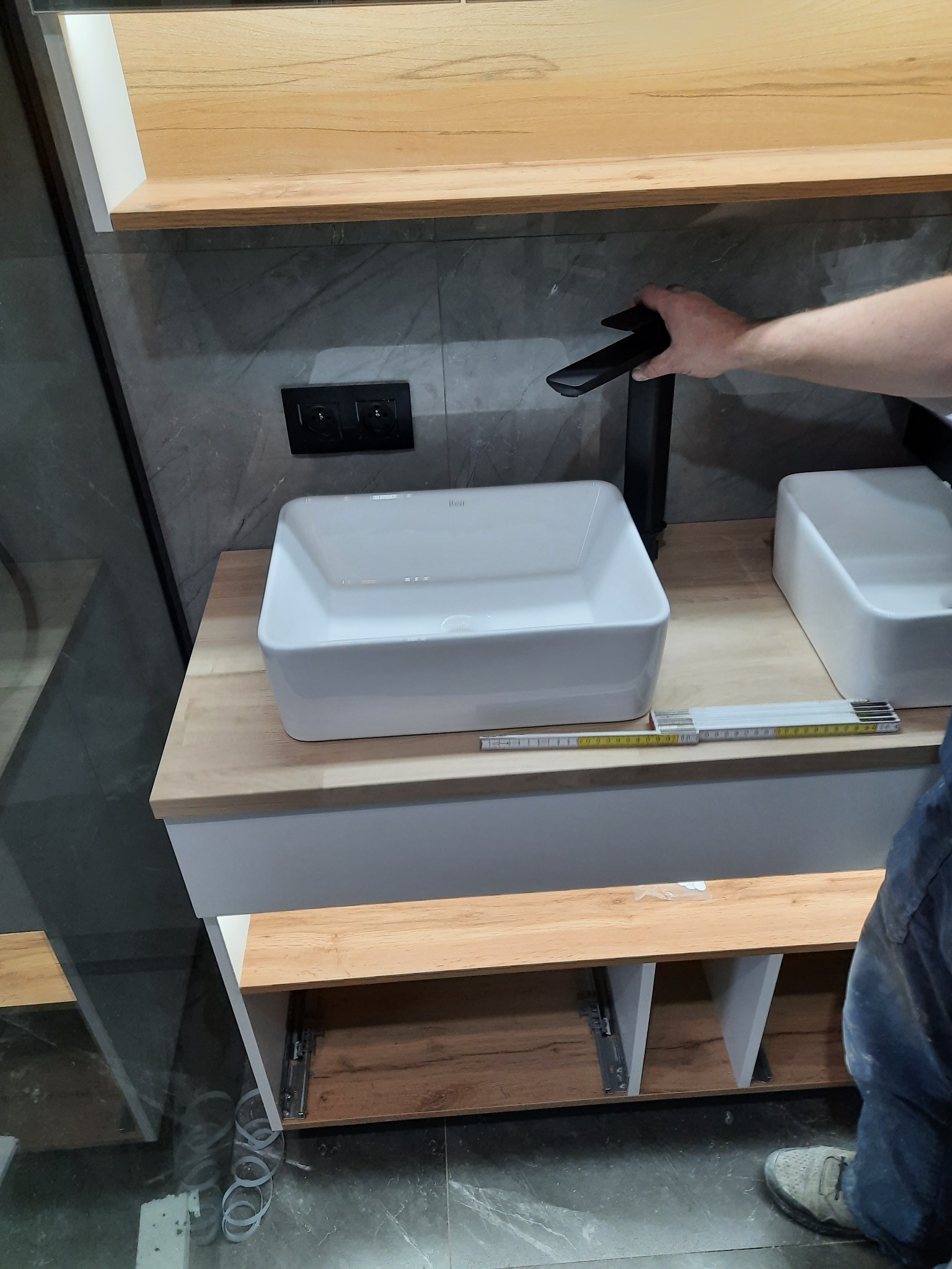 Montaż czarnej baterii umywalkowej na blacie z drewnopodobnym dekorem, obok biała umywalka nablatowa i złożona miarka stolarska, szare płytki na ścianie.