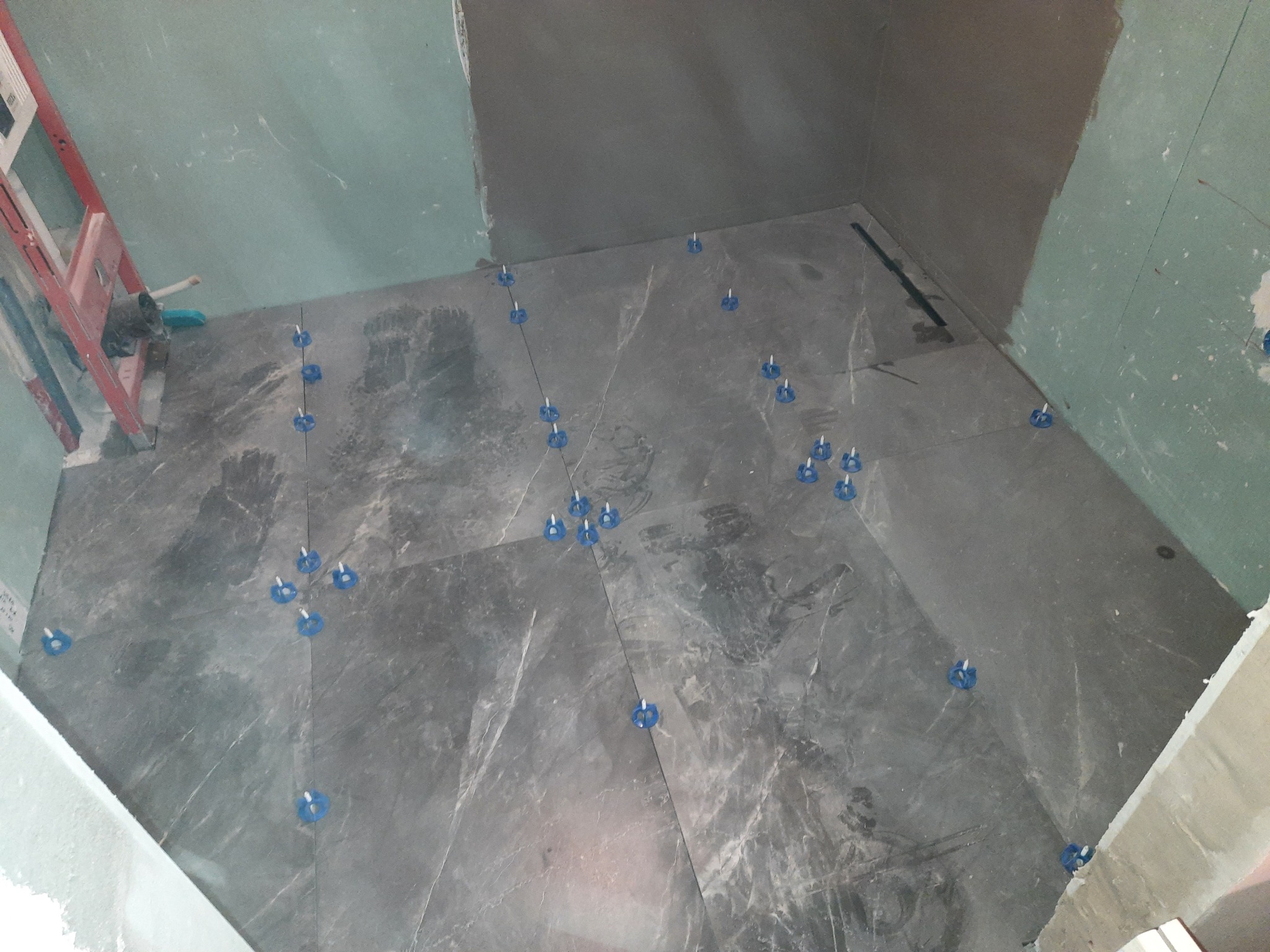 Układanie szarych płytek podłogowych z niebieskimi klipsami poziomującymi w narożnej części pomieszczenia z surowymi ścianami przygotowanymi do dalszych prac.