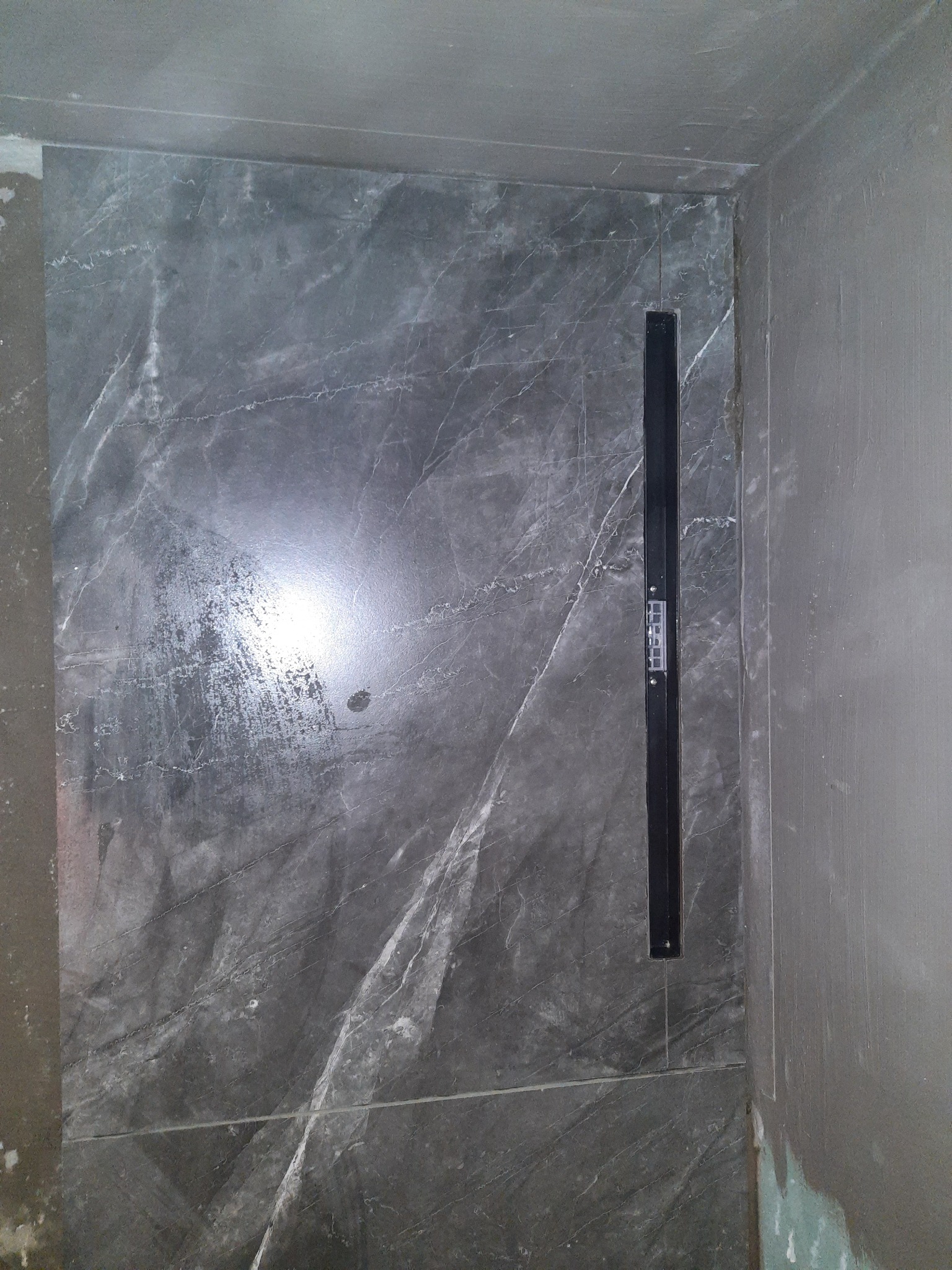 Ściana prysznica wyłożona szarymi płytkami imitującymi marmur z zamontowanym liniowym odpływem; widoczne ślady zaprawy.