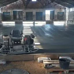 Wnętrze hali z wylewką betonową, na której stoi zacieraczka mechaniczna do betonu. Widoczne worki z zaprawą Tybet Hard i wiadro na palecie. W tle okna w starym budynku.