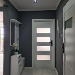 Szare wnętrze przedpokoju z białymi drzwiami z poziomymi przeszkleniami, białą szafką na buty z lustrem i wieszakami oraz jasną podłogą z paneli.