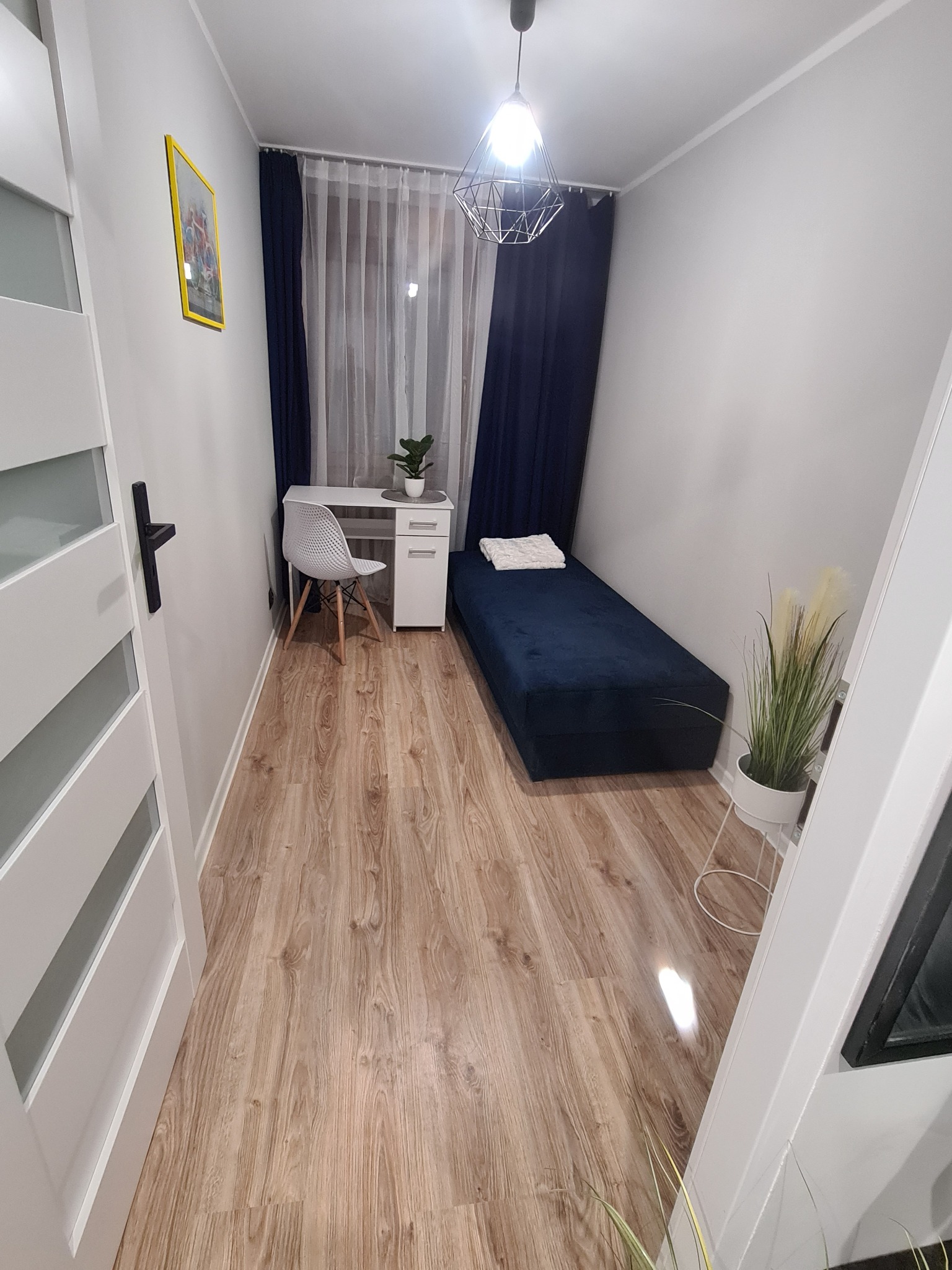 Wnętrze pokoju z panelem podłogowym w odcieniach drewna,  ciemnoniebieską sofą, białym biurkiem z krzesłem, i dekoracjami roślinnymi, oświetlone nowoczesną lampą wiszącą.