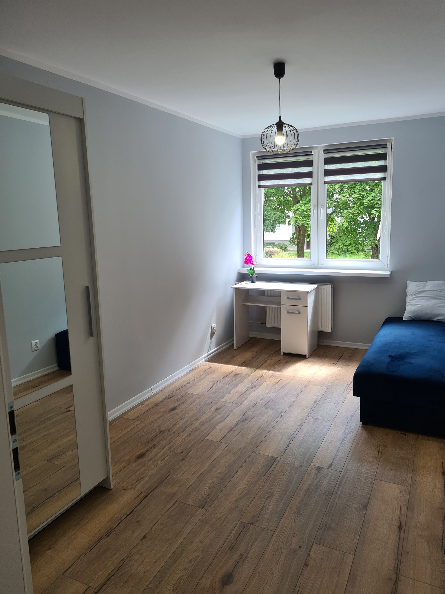 Pokój z drewnianą podłogą ułożoną z paneli, biurkiem przy oknie, szafą z lustrem i niebieską sofą.