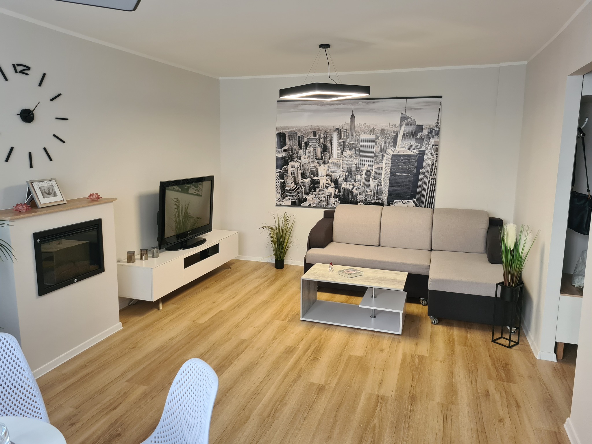 Salon z panelami podłogowymi w odcieniach drewna, szara sofa narożna, biała szafka RTV i kominek elektryczny, na ścianie czarno-biała panorama Nowego Jorku.