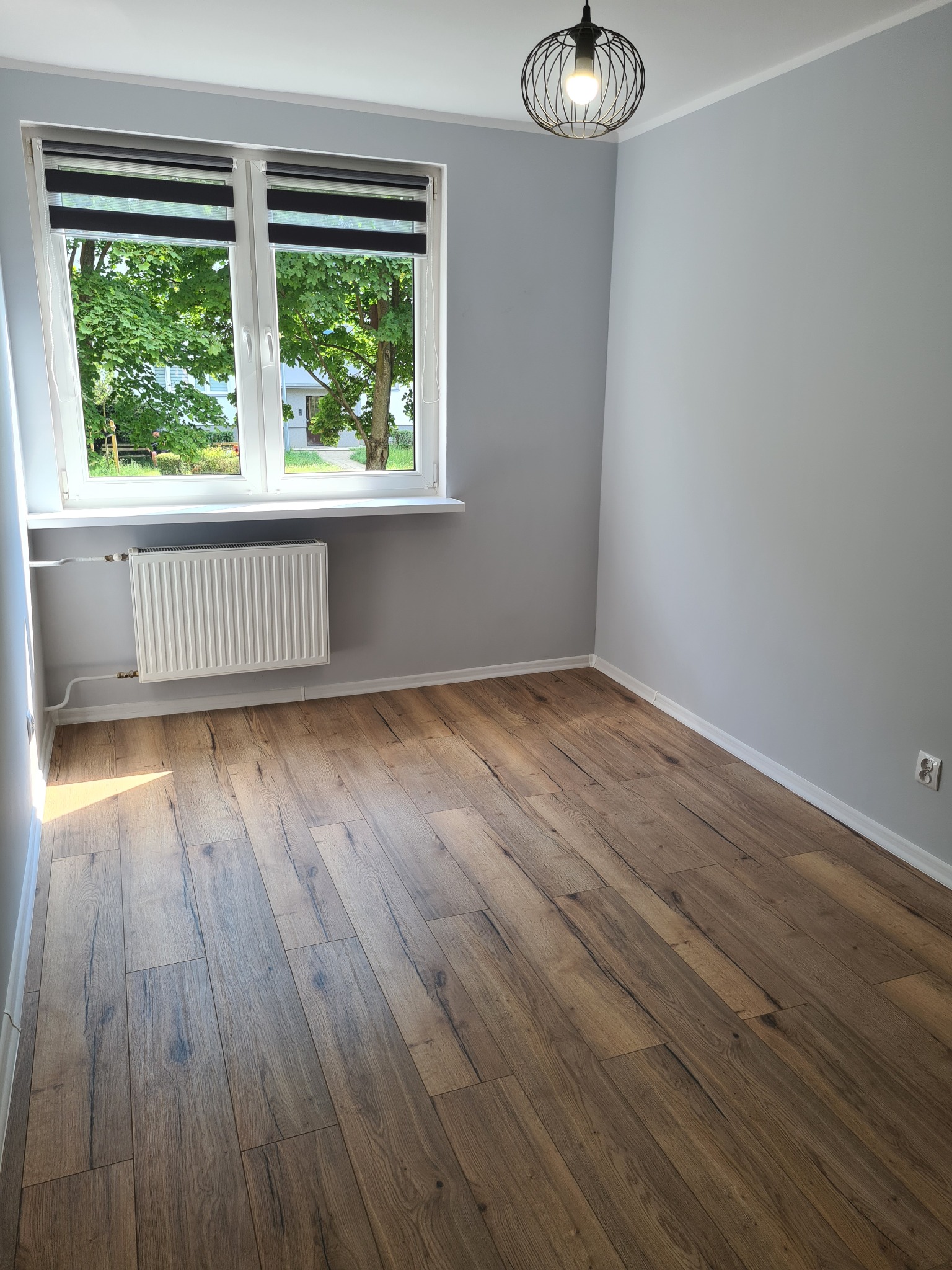 Pusty pokój z drewnianą podłogą ułożoną w jodełkę, szare ściany, białe okno z roletami dzień-noc i widokiem na zieleń, oraz designerska lampa wisząca.