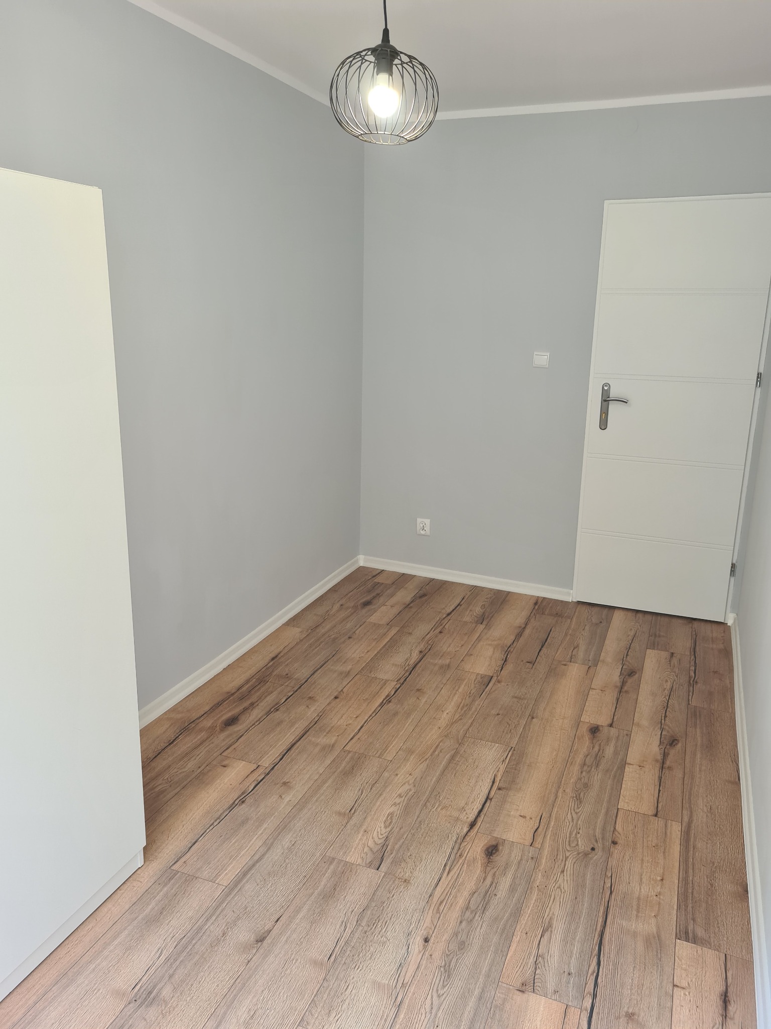 Jasne panele podłogowe ułożone w pustym pokoju z szarymi ścianami, białą szafą i drzwiami oraz wiszącą lampą drucianą.