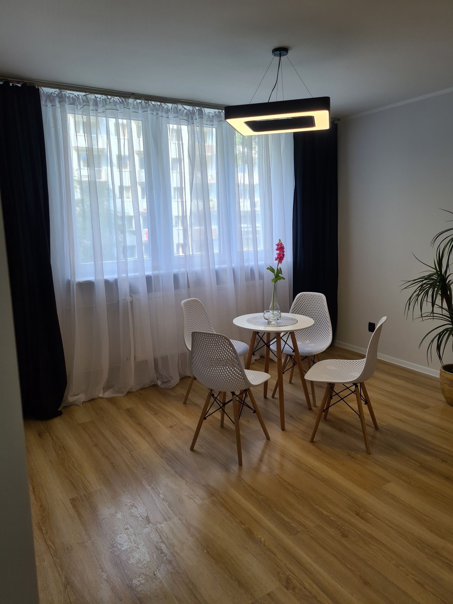 Jasne panele podłogowe w pokoju z minimalistycznym wystrojem, okno z białymi firanami i czarnymi zasłonami, okrągły stolik z czterema krzesłami i nowoczesna lampa sufitowa.