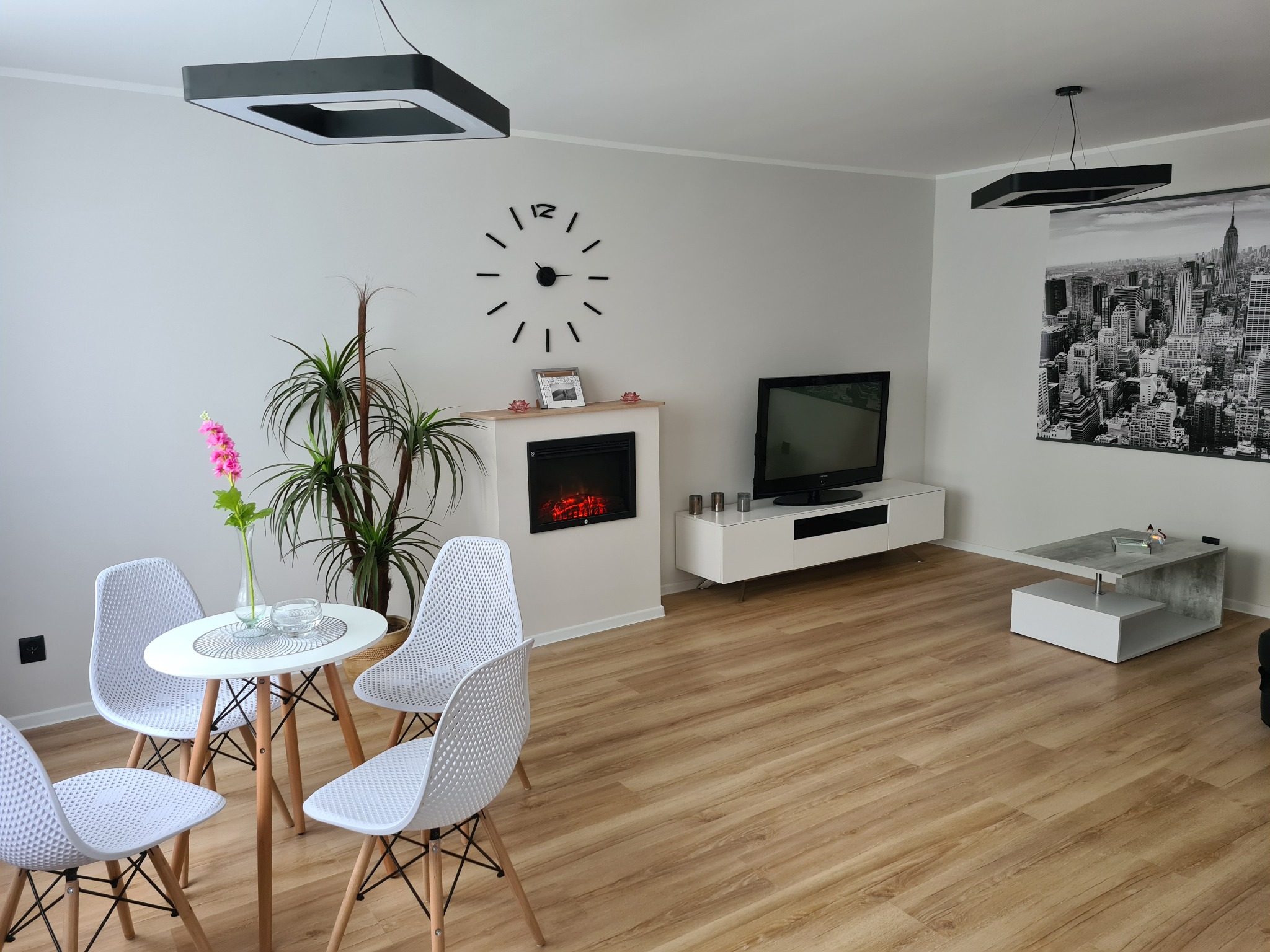 Jasne wnętrze salonu z drewnianą podłogą, okrągłym stołem z białymi krzesłami, elektrycznym kominkiem i minimalistycznymi lampami sufitowymi.