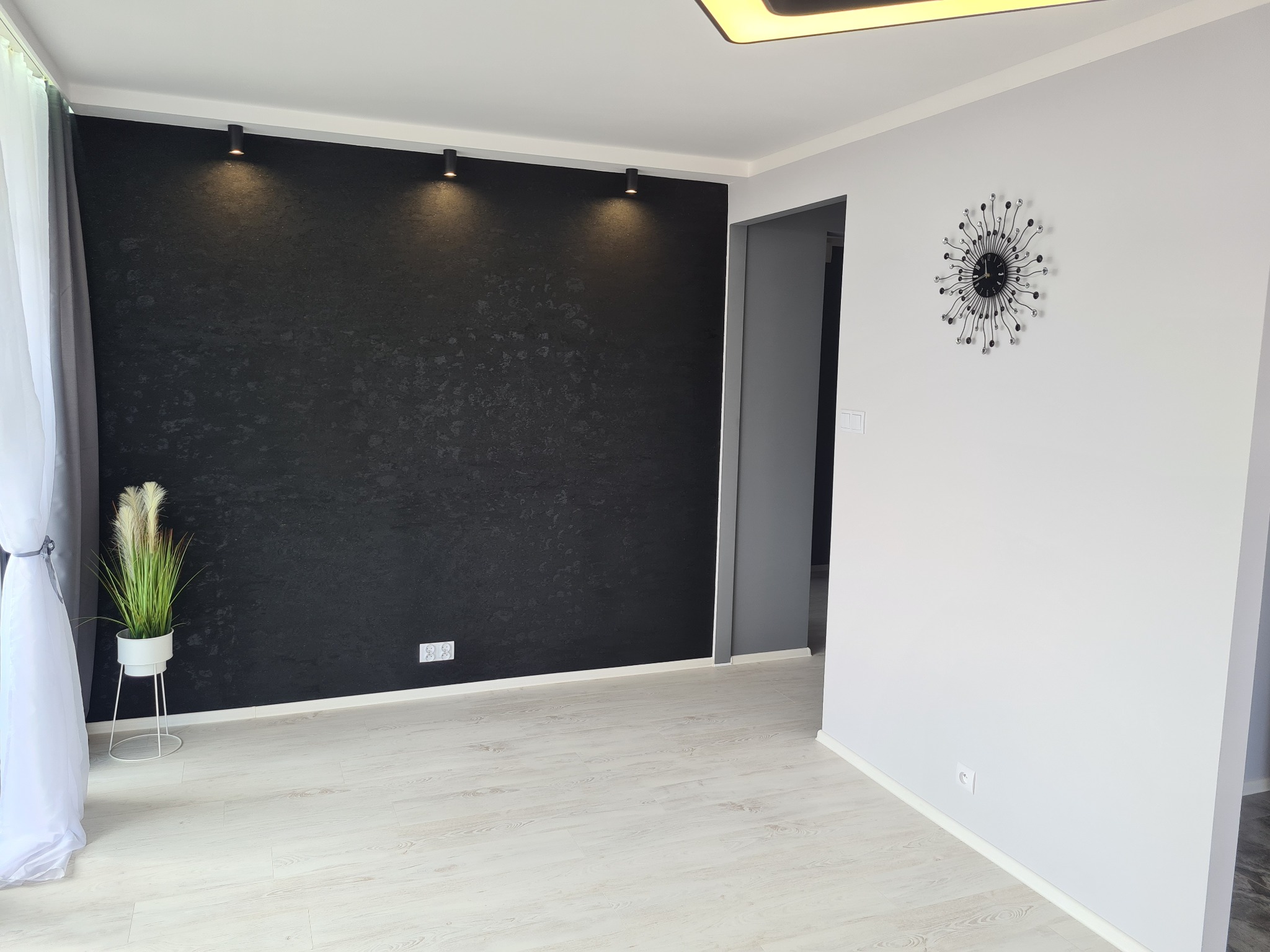 Jasne panele podłogowe w nowoczesnym salonie z czarną ścianą akcentującą, oświetloną punktowo, oraz dekoracyjnym zegarem na szarej ścianie
