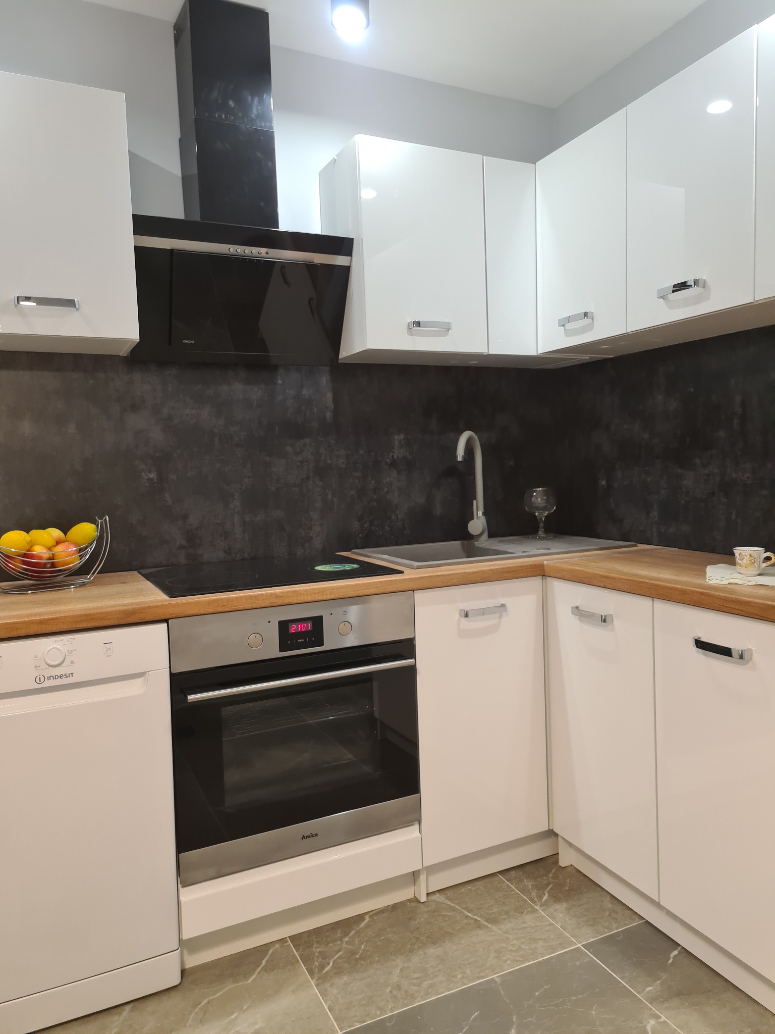 Nowoczesna kuchnia w kształcie litery L z białymi, błyszczącymi szafkami, drewnianym blatem, czarnym panelem nad blatem, zintegrowanym piekarnikiem i płytą indukcyjną oraz metalowym koszem z owocami.