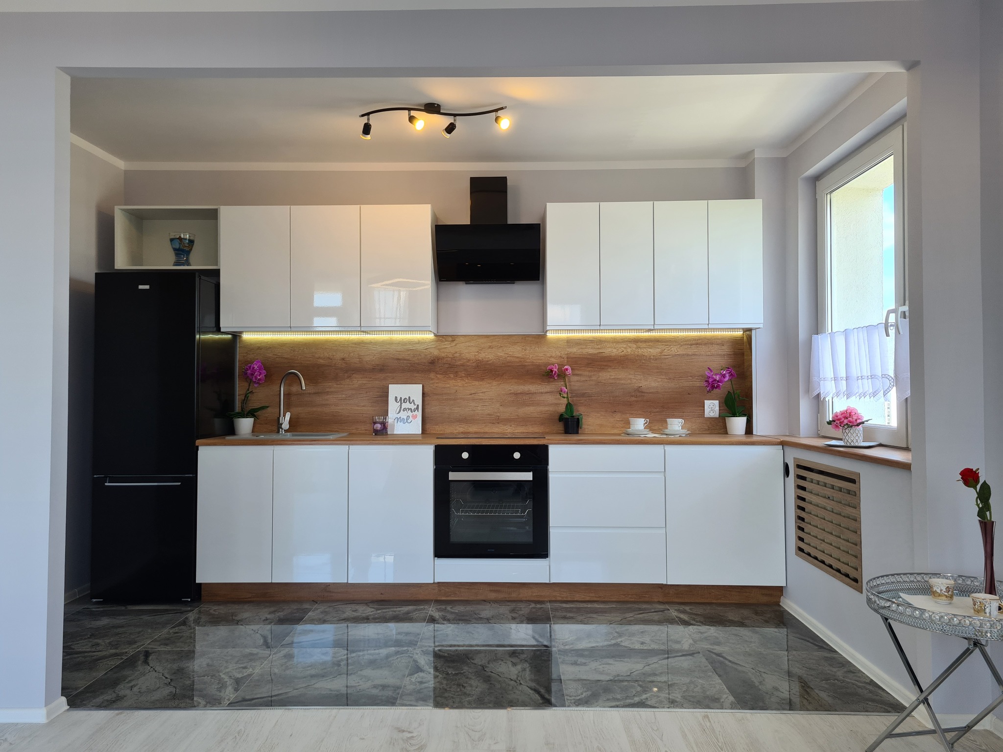 Nowoczesna kuchnia z białymi, lakierowanymi szafkami, drewnianym blatem i podświetleniem LED, czarną lodówką i piekarnikiem oraz marmurową podłogą.