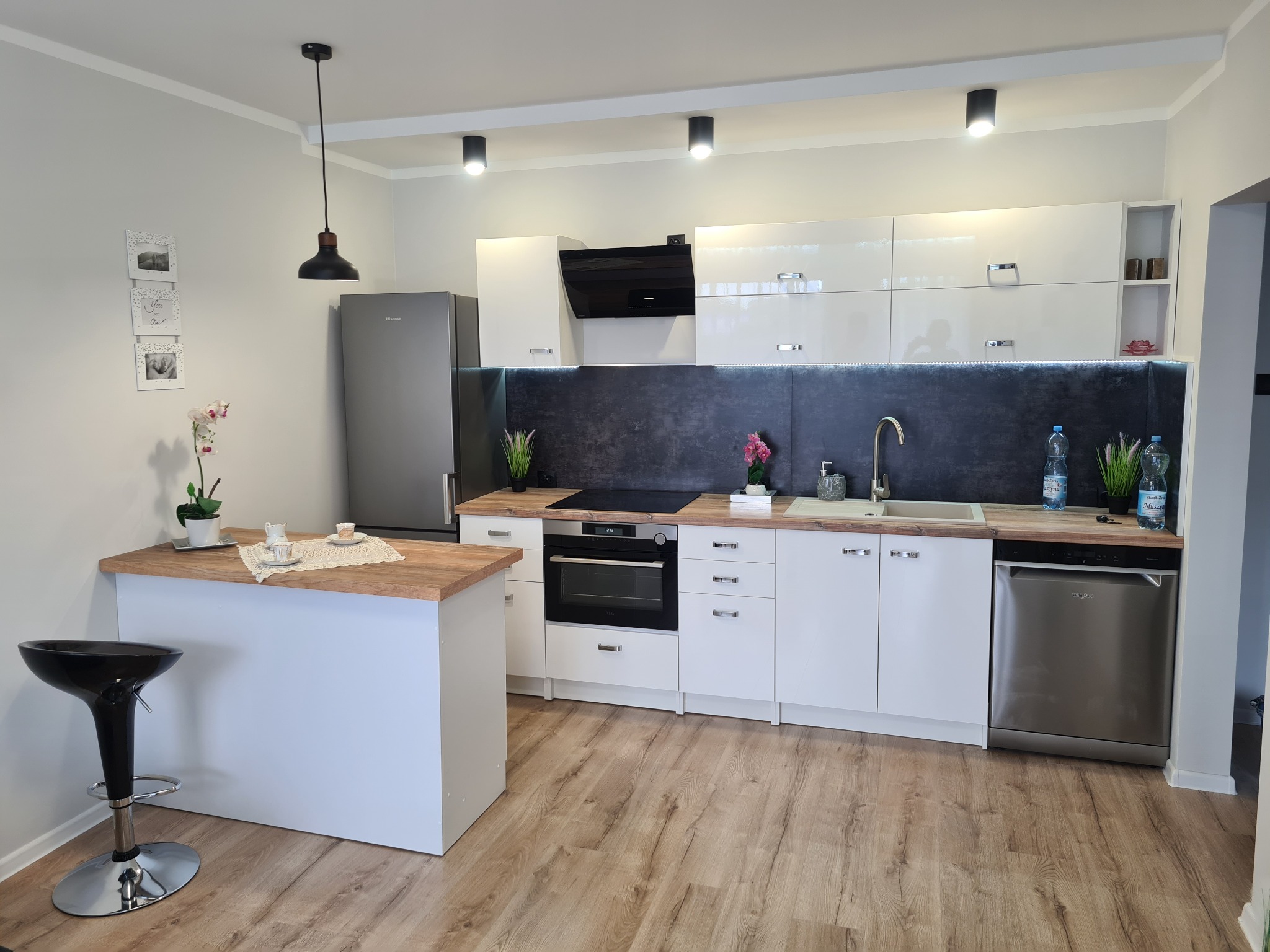 Nowoczesna kuchnia z białymi błyszczącymi szafkami, drewnianym blatem, czarnym okapem i podłogą imitującą drewno. Na blacie widoczne dekoracje, w tle srebrna lodówka.