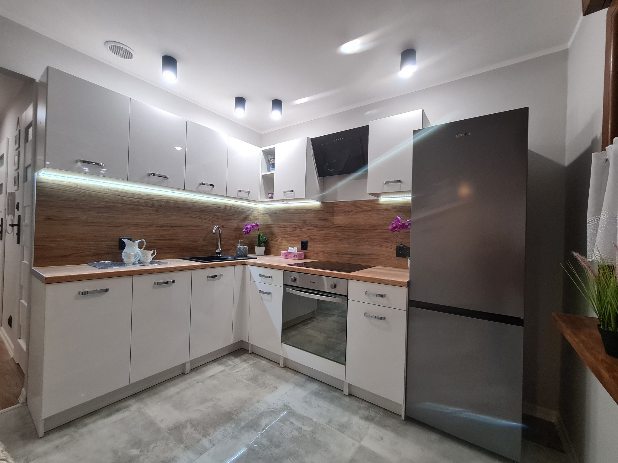 Nowoczesna kuchnia w odcieniach bieli i drewna z podświetleniem LED, srebrna lodówka Gorenje, czarny okap i szary blat, całość dopełniona dekoracjami.