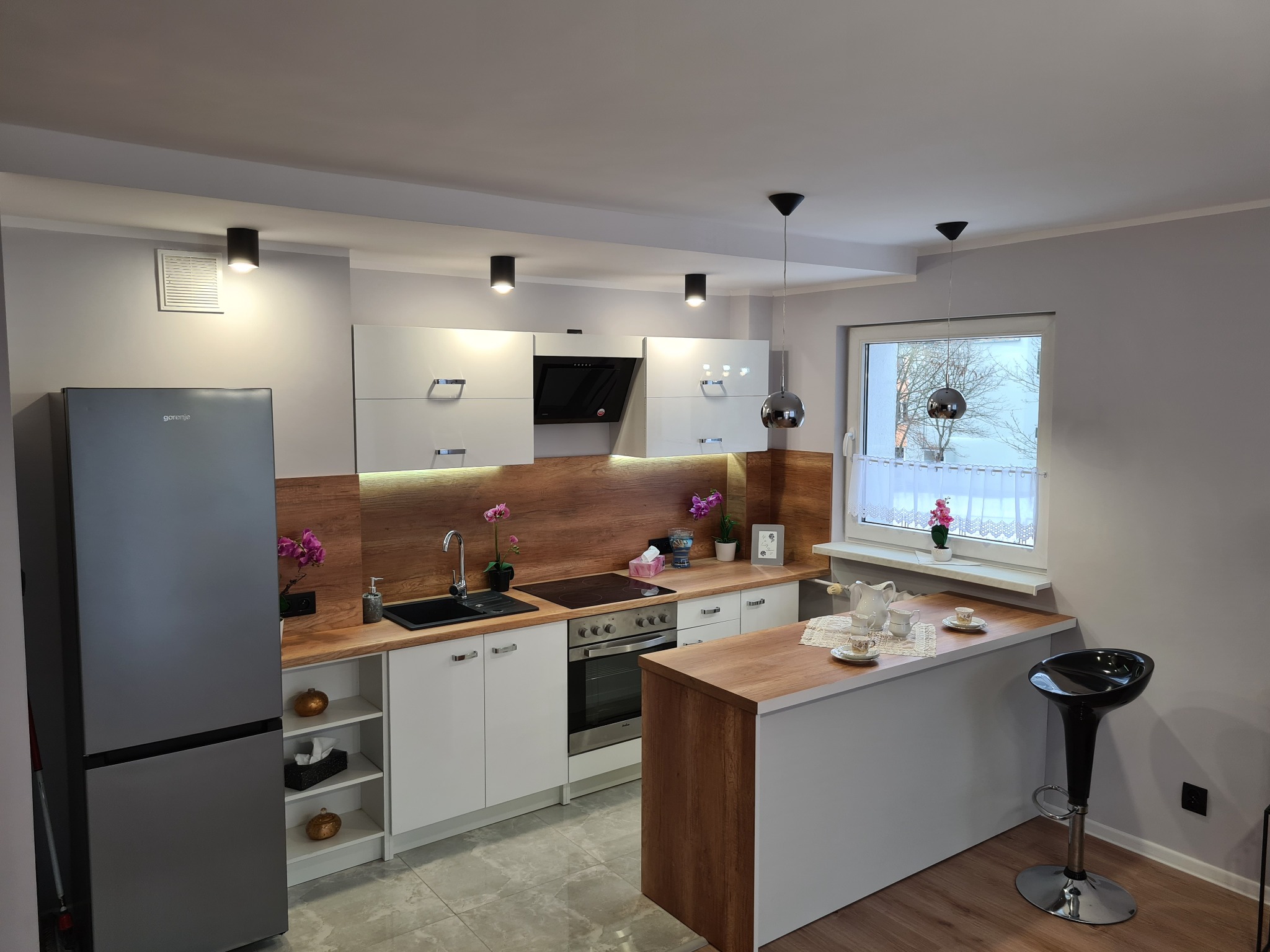 Nowoczesna kuchnia z białymi szafkami, drewnianym blatem i wyspą, oświetlona punktowymi lampami sufitowymi i dekorowana kwiatami w doniczkach.