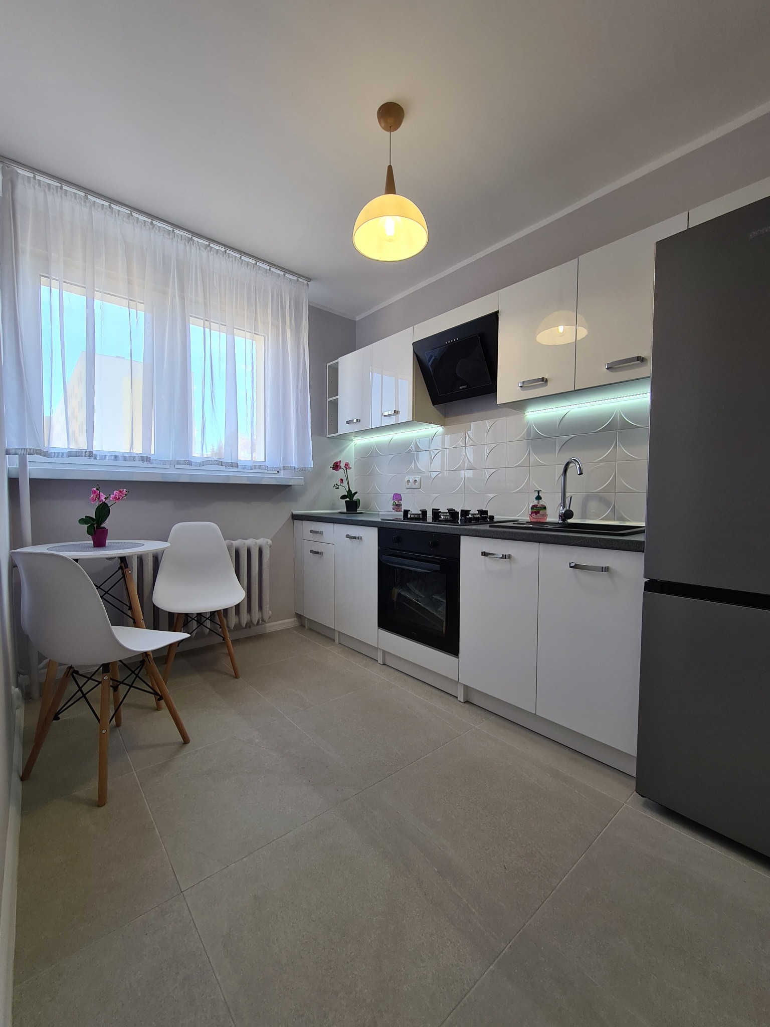 Nowoczesna kuchnia z białymi szafkami, czarnym sprzętem AGD, podświetleniem LED pod szafkami wiszącymi i stołem z dwoma krzesłami przy oknie z firanką.