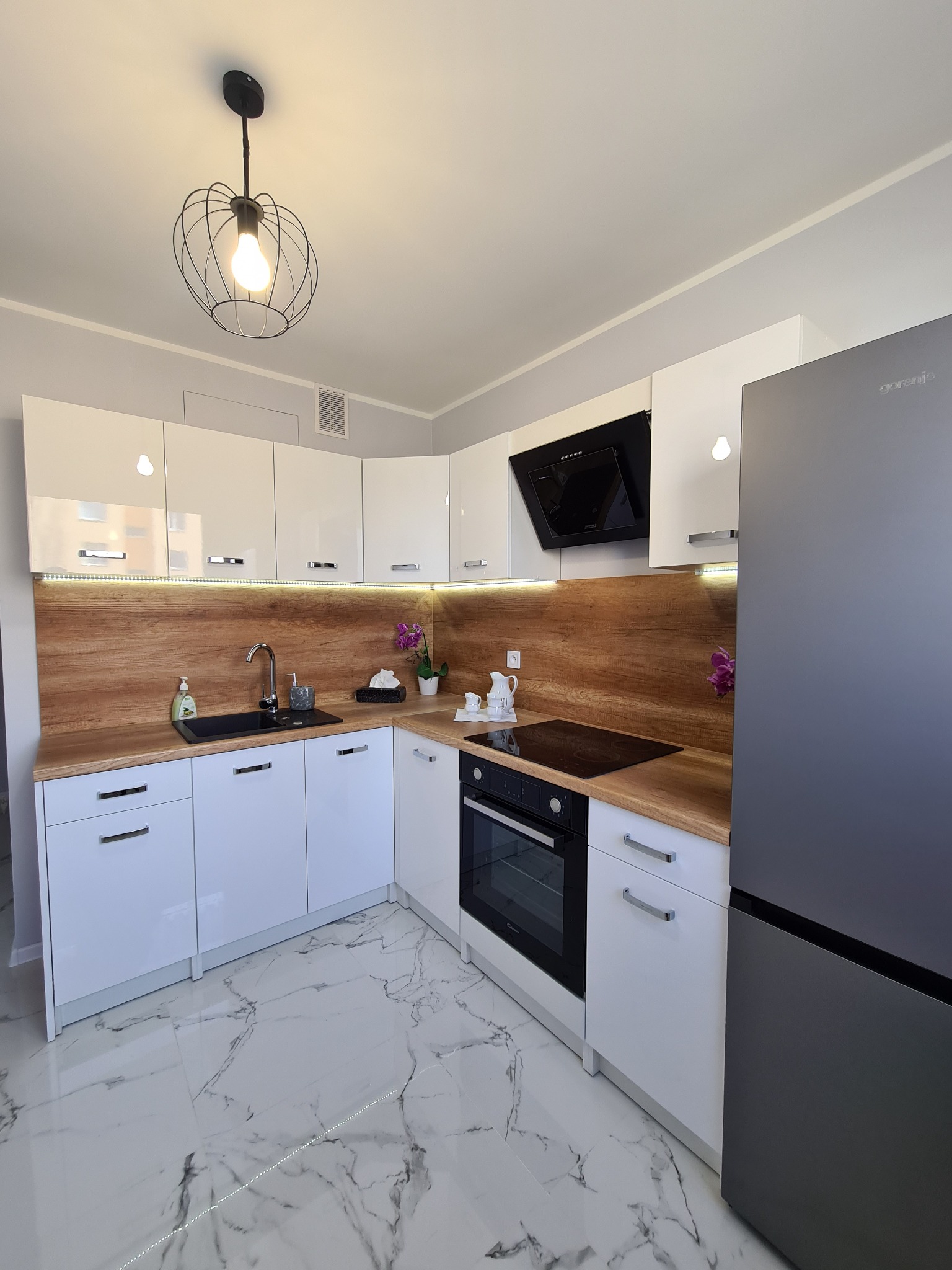 Nowoczesna kuchnia z białymi, lakierowanymi szafkami, drewnianym blatem i marmurową podłogą, oświetlona lampą wiszącą w stylu industrialnym. Widoczny czarny zlew, płyta indukcyjna i szary lodówka.