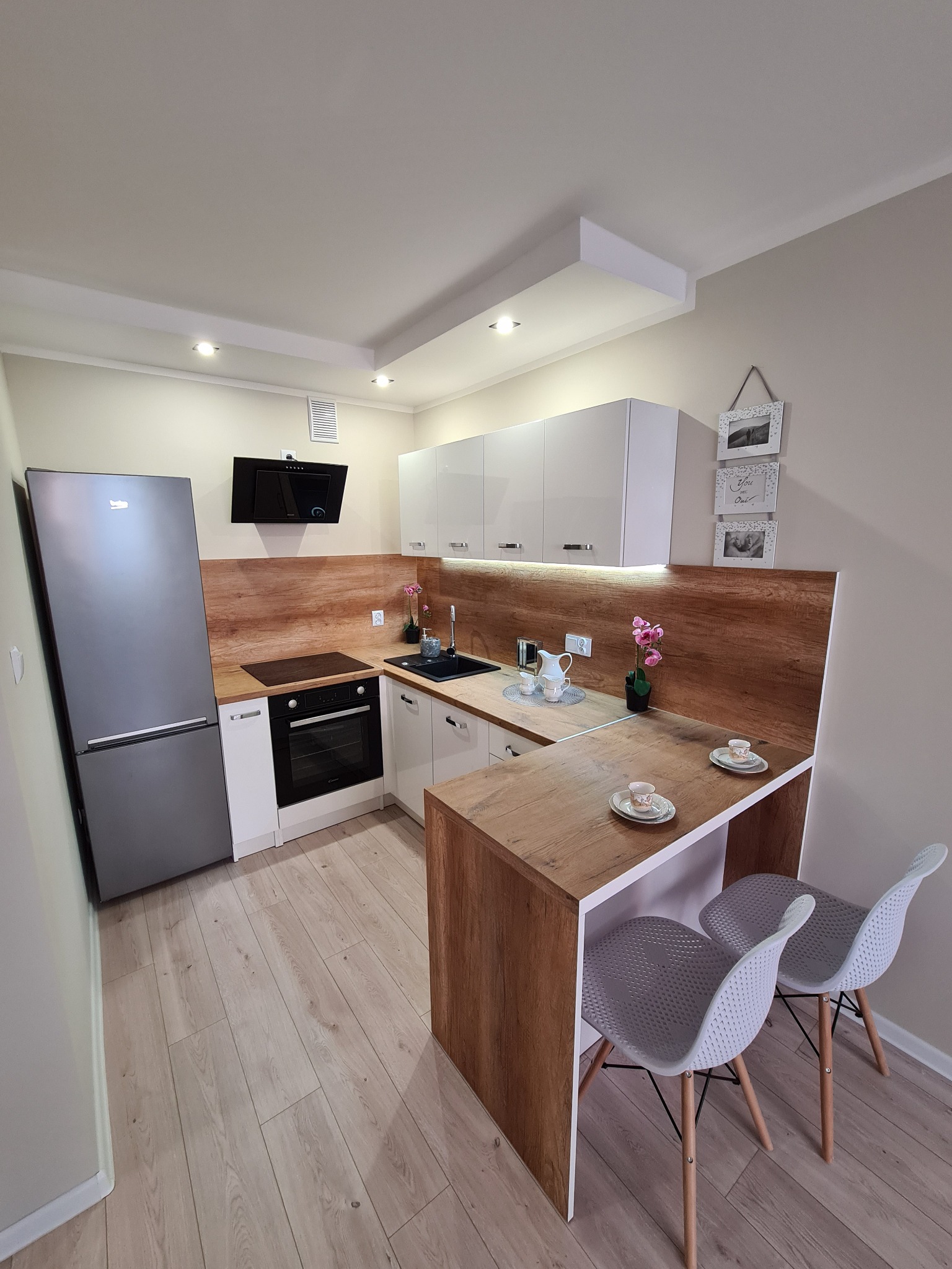 Nowoczesna kuchnia z drewnianym blatem, białymi szafkami i szarym sprzętem AGD, uzupełniona o dwie filiżanki na blacie i minimalistyczne krzesła.