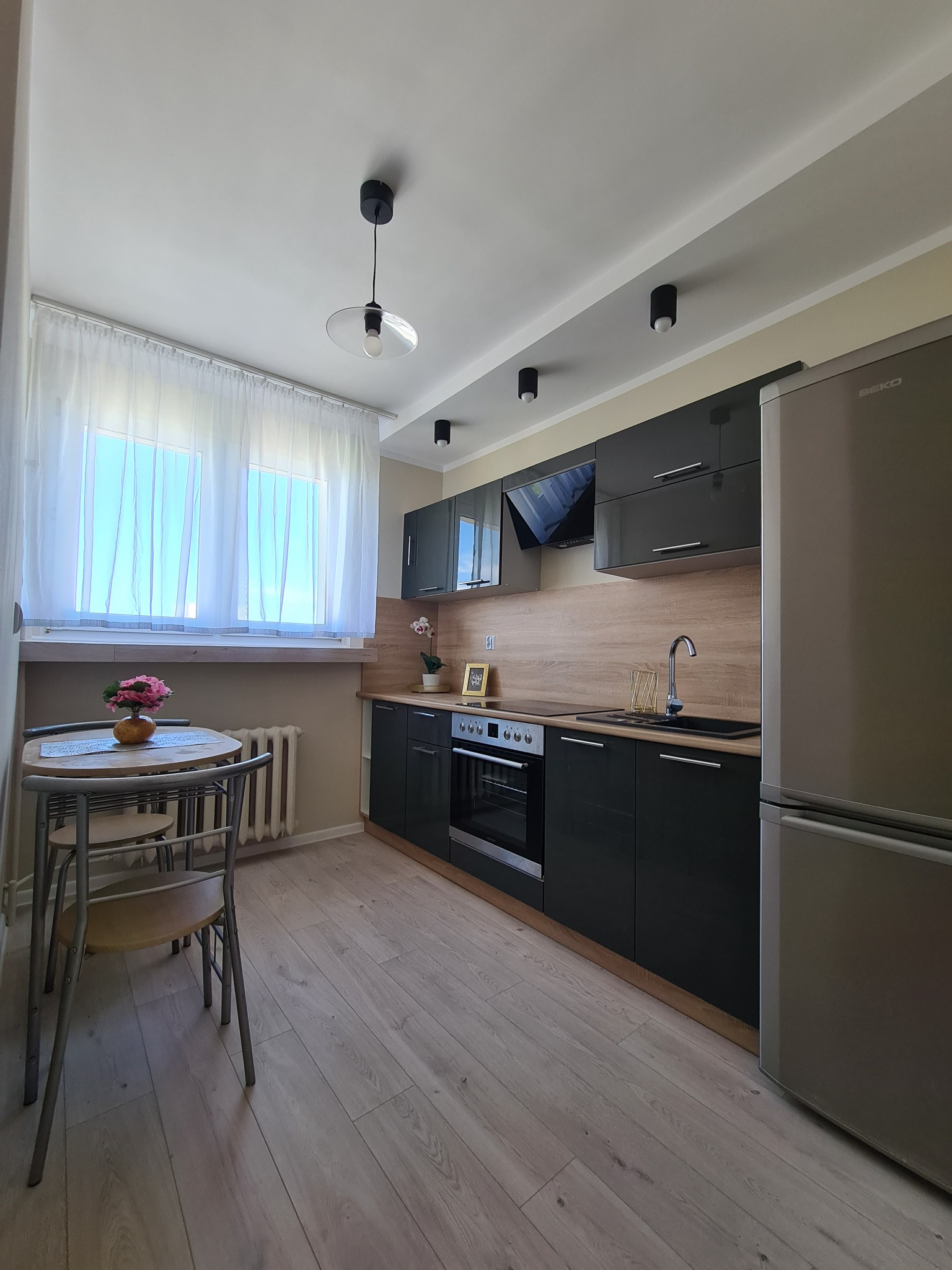 Nowoczesna kuchnia z czarnymi, lakierowanymi szafkami, drewnianym blatem i jasną podłogą. Widoczny stół z kwiatami przy oknie.