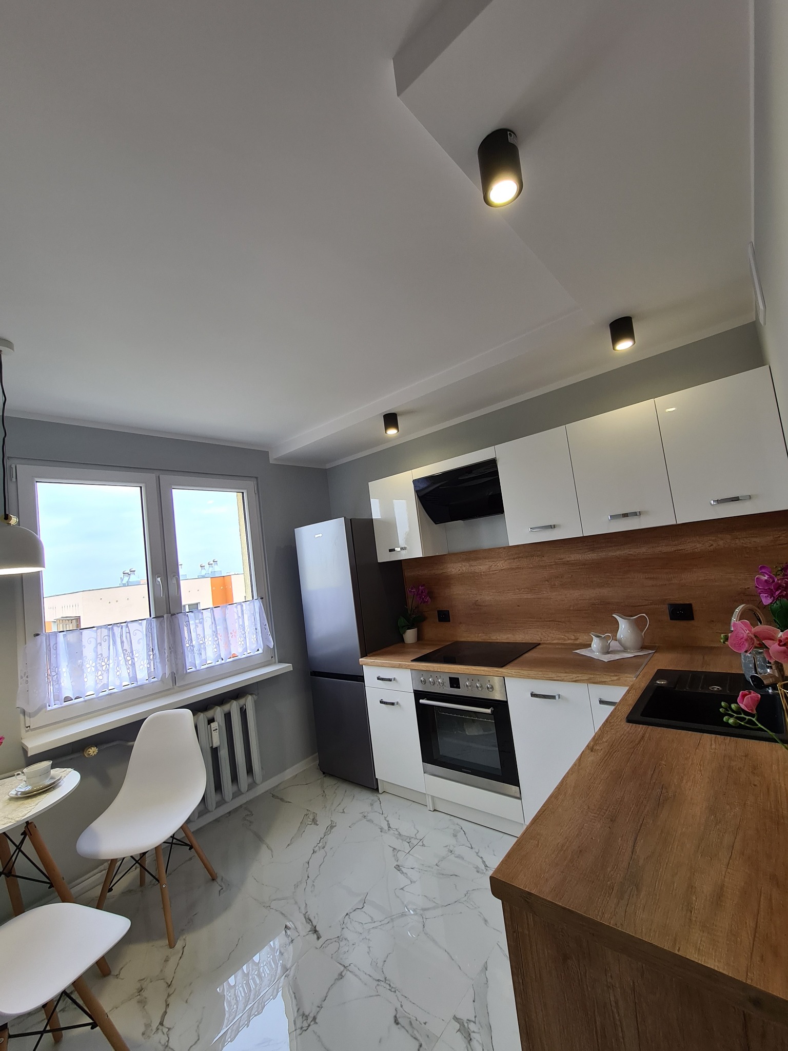 Nowoczesna kuchnia z białymi szafkami, drewnianym blatem i marmurową podłogą, widok z góry na stół z krzesłami przy oknie.