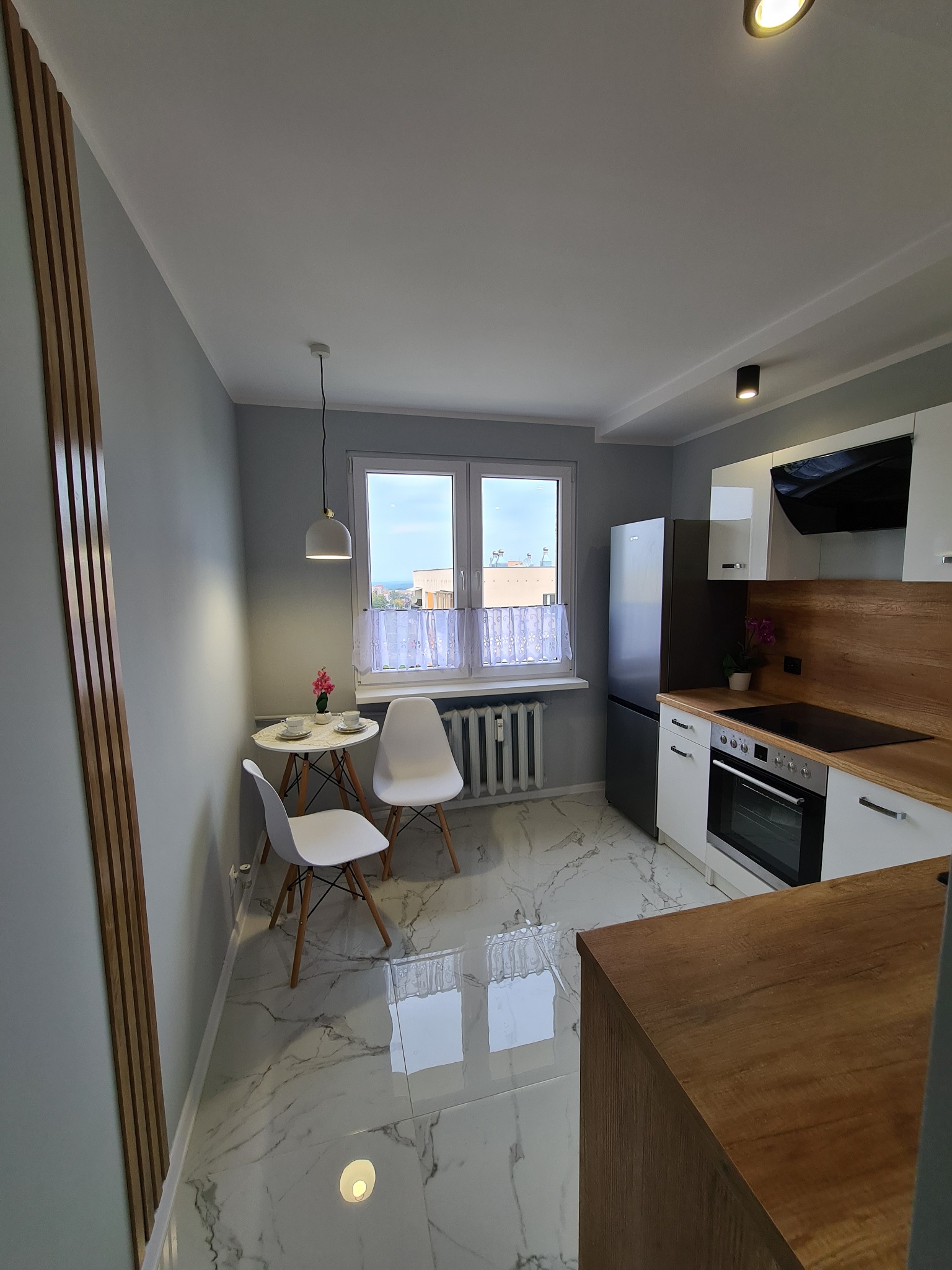 Jasna kuchnia z marmurową podłogą, drewnianymi akcentami na ścianie i blacie, białymi meblami, okrągłym stolikiem kawowym z dwiema filiżankami i kwiatkiem, oraz białymi krzesłami na drewnianych...