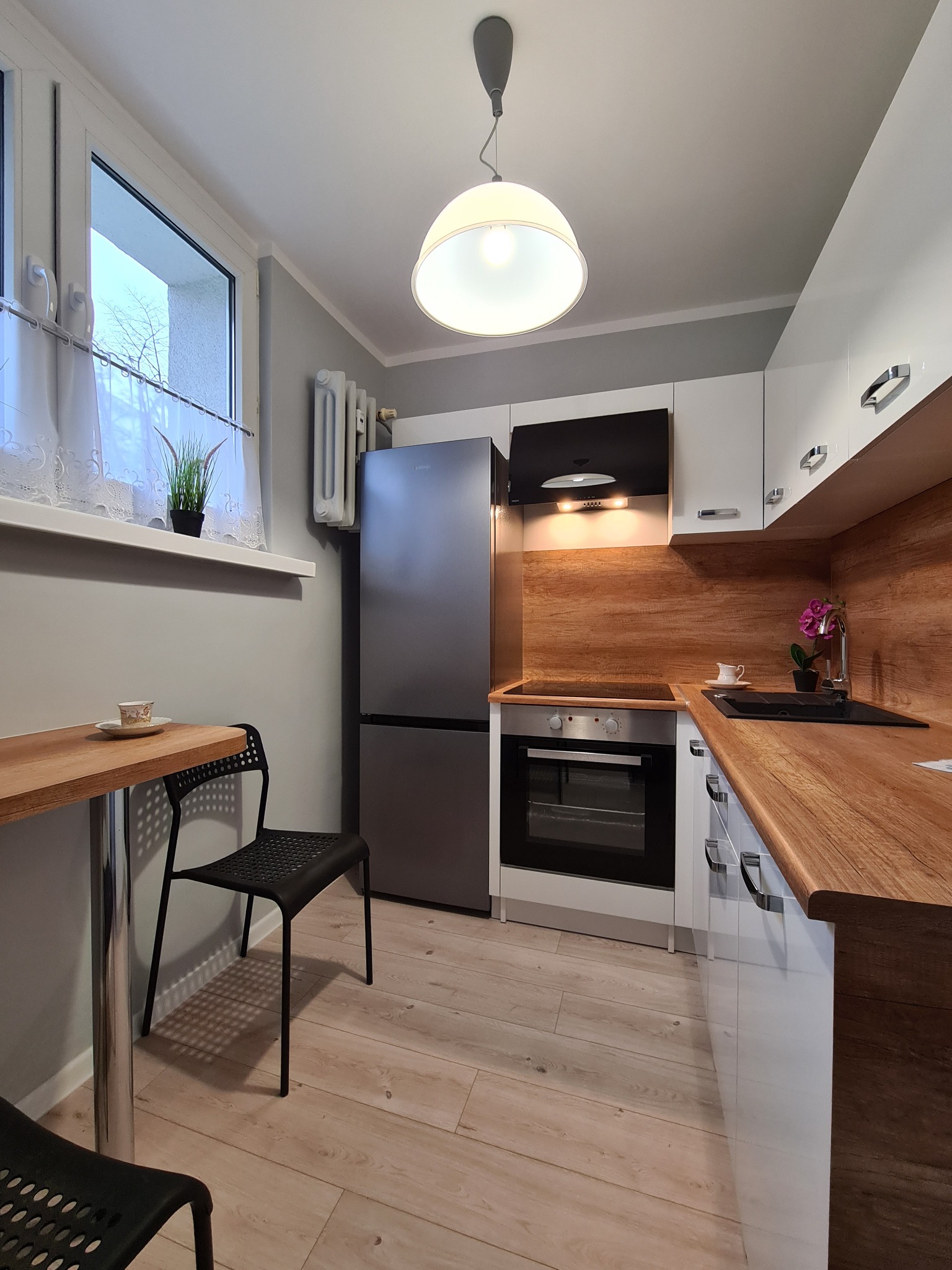 Nowoczesna kuchnia z białymi szafkami, drewnianym blatem i panelem ściennym, szara lodówka, czarne krzesła i stół, podłoga z paneli imitujących drewno, oświetlenie sufitowe, filiżanka na blacie.