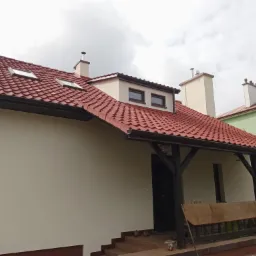 Dom z nową, czerwoną dachówką, ocieploną elewacją w kolorze kremowym i drewnianą werandą z czarnymi słupami, widok z dołu na pochmurne niebo.