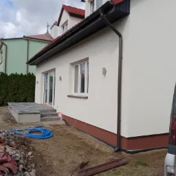 Świeżo pomalowana elewacja domu jednorodzinnego w kolorze kremowym, z brązowym cokołem, widoczne rynny i fragment nowo wybudowanego tarasu z kostki brukowej, obok sterta gruzu i zaparkowany...