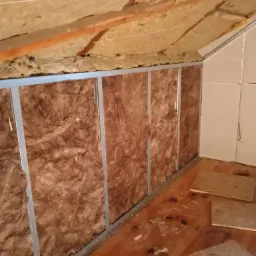 Poddasze w trakcie izolacji: wełna mineralna w metalowej konstrukcji, drewniane belki stropowe i częściowo zamontowane płyty kartonowo-gipsowe na ścianie szczytowej, widoczne fragmenty podłogi...