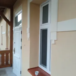 Jasna elewacja budynku z białymi detalami architektonicznymi, okno z brązowym parapetem i filiżanką, białe drzwi wejściowe z przeszklonym nadświetlem.