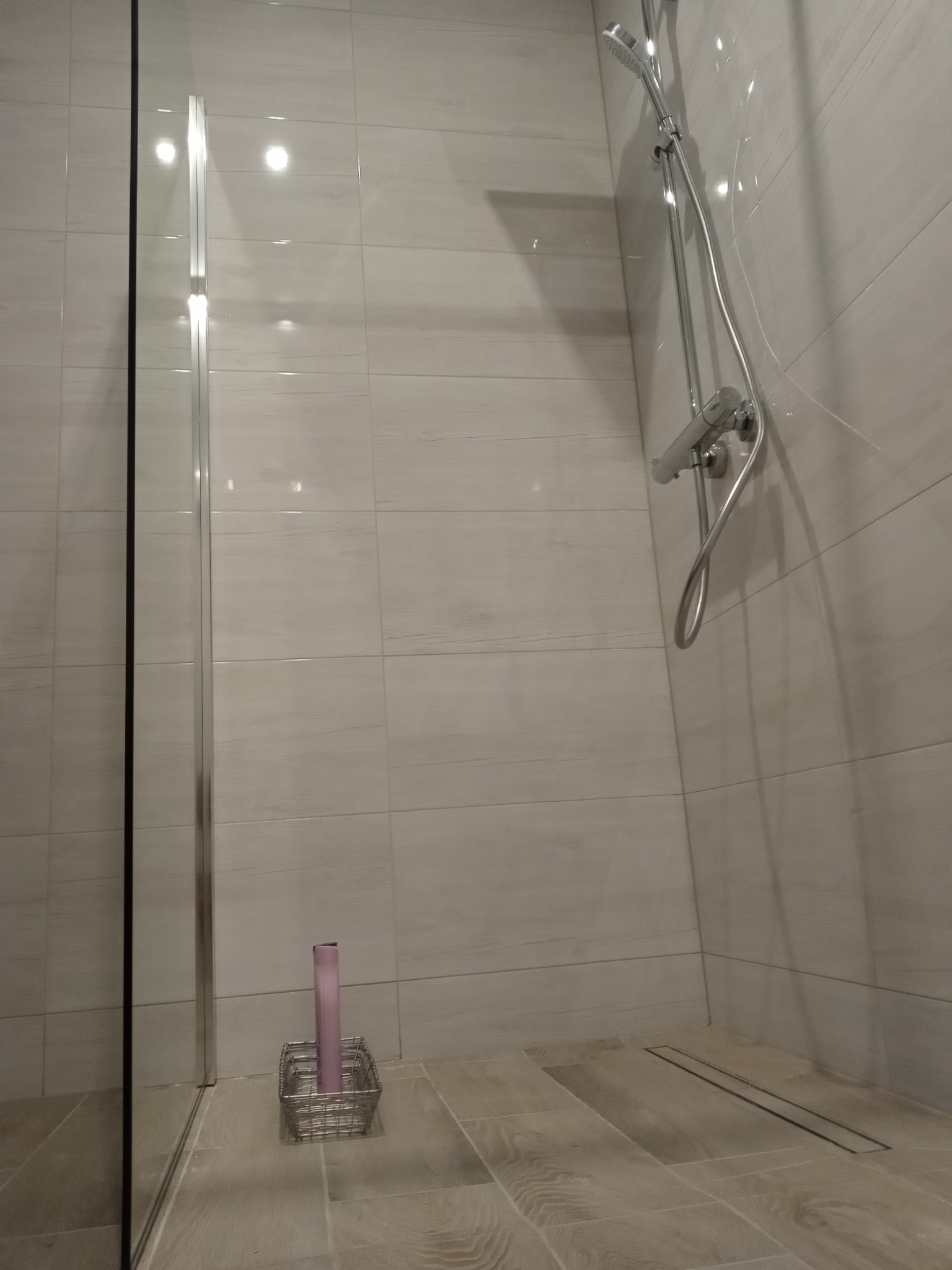 Nowoczesna kabina prysznicowa z przeszklonymi drzwiami, wyłożona jasnymi płytkami imitującymi drewno, z chromowaną armaturą i odpływem liniowym w podłodze; w rogu widoczny koszyk na kosmetyki...