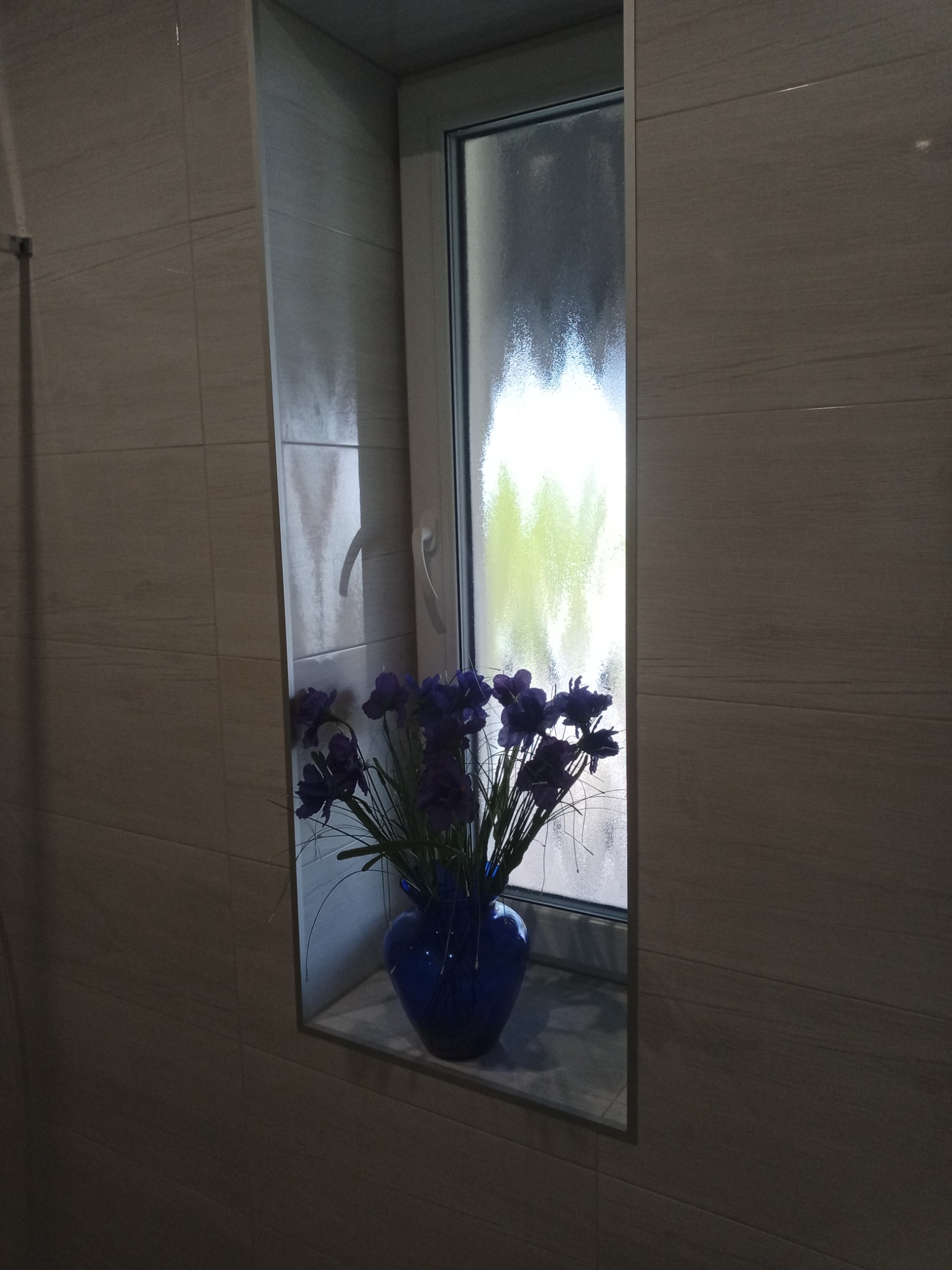 Wnęka okienna w łazience wyłożona płytkami imitującymi drewno, z niebieskim wazonem z fioletowymi kwiatami na parapecie.