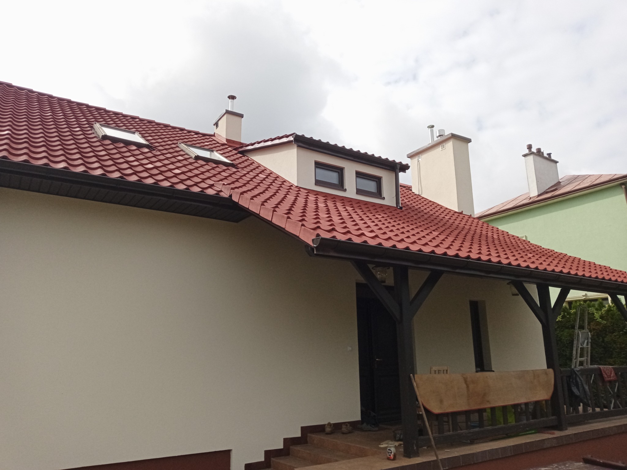 Dom z nową, czerwoną dachówką, ocieploną elewacją w kolorze kremowym i drewnianą werandą z czarnymi słupami, widok z dołu na pochmurne niebo.