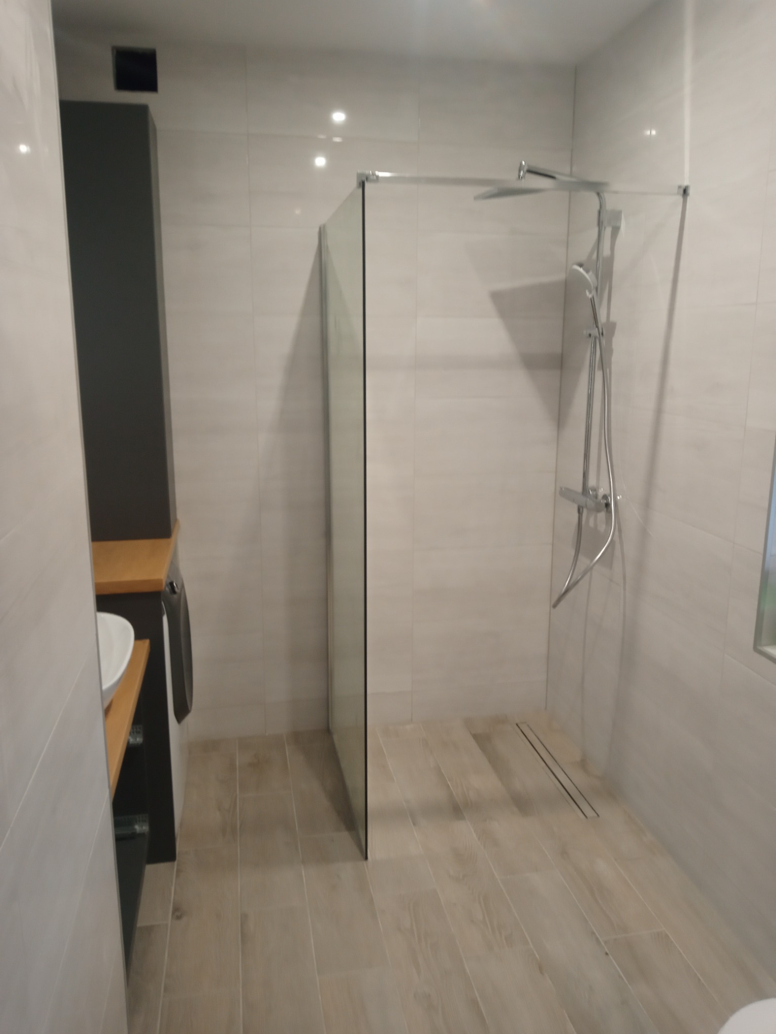 Nowoczesna łazienka z kabiną prysznicową typu walk-in, wykończona płytkami w odcieniach beżu i szarości, z widoczną armaturą chromowaną i minimalistycznym odpływem liniowym w podłodze.