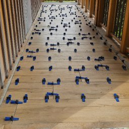 Rembau Remigiusz Leśnik - Układanie desek tarasowych na balkonie z widokiem na zieleń, z użyciem niebieskich klipsów dystansowych i miarki. Drewniana balustrada i drzwi w tle.