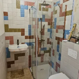 Wykończona łazienka z kabiną prysznicową, umywalką i podwieszaną toaletą. Ściany wyłożone mozaiką z kolorowych płytek, podłoga z kamiennych płyt. Elementy wykończeniowe w kolorze miedzi.