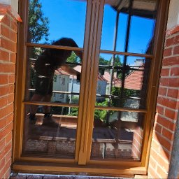 Rembau Remigiusz Leśnik - Nowe okno drewniane z szybami refleksyjnymi, zamontowane w murze z czerwonej cegły, widok z zewnątrz, fragment rusztowania, pracownik w ubraniu roboczym.