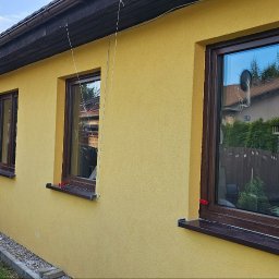 Rembau Remigiusz Leśnik - Żółta elewacja domu z trzema brązowymi oknami. W oknach odbija się zieleń drzew i niebo. Obok okien widoczna brązowa rynna. Na trawie leżą płyty ociepleniowe.