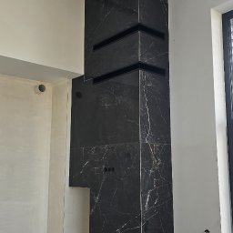 Rembau Remigiusz Leśnik - Wykończenie kominka czarnymi płytkami imitującymi marmur z widocznymi liniowymi kratkami wentylacyjnymi. Prace wykończeniowe w surowym wnętrzu.