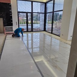 Rembau Remigiusz Leśnik - Glazurnik układa błyszczące, białe płytki podłogowe z delikatnym wzorem w przestronnym wnętrzu z dużymi oknami. Widoczne listwy przypodłogowe i narzędzia.