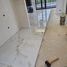 Rembau Remigiusz Leśnik - Układanie płytek podłogowych w przestronnym wnętrzu. Widoczne narzędzia glazurnicze i częściowo ułożone, białe płytki z marmurkowym wzorem. W tle okno i pracujący glazurnik.