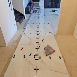 Rembau Remigiusz Leśnik - Układanie jasnych, marmurkowych płytek podłogowych z użyciem klipsów poziomujących. Dwóch glazurników pracuje w tle, widoczna część schodów i okna.