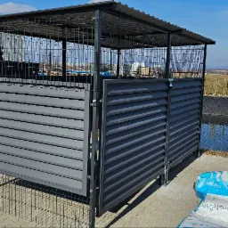 Szara, metalowa wiata śmietnikowa z ażurową górą i poziomymi lamelami, na kostce brukowej, w tle krajobraz. Widoczne worki z tworzywem.