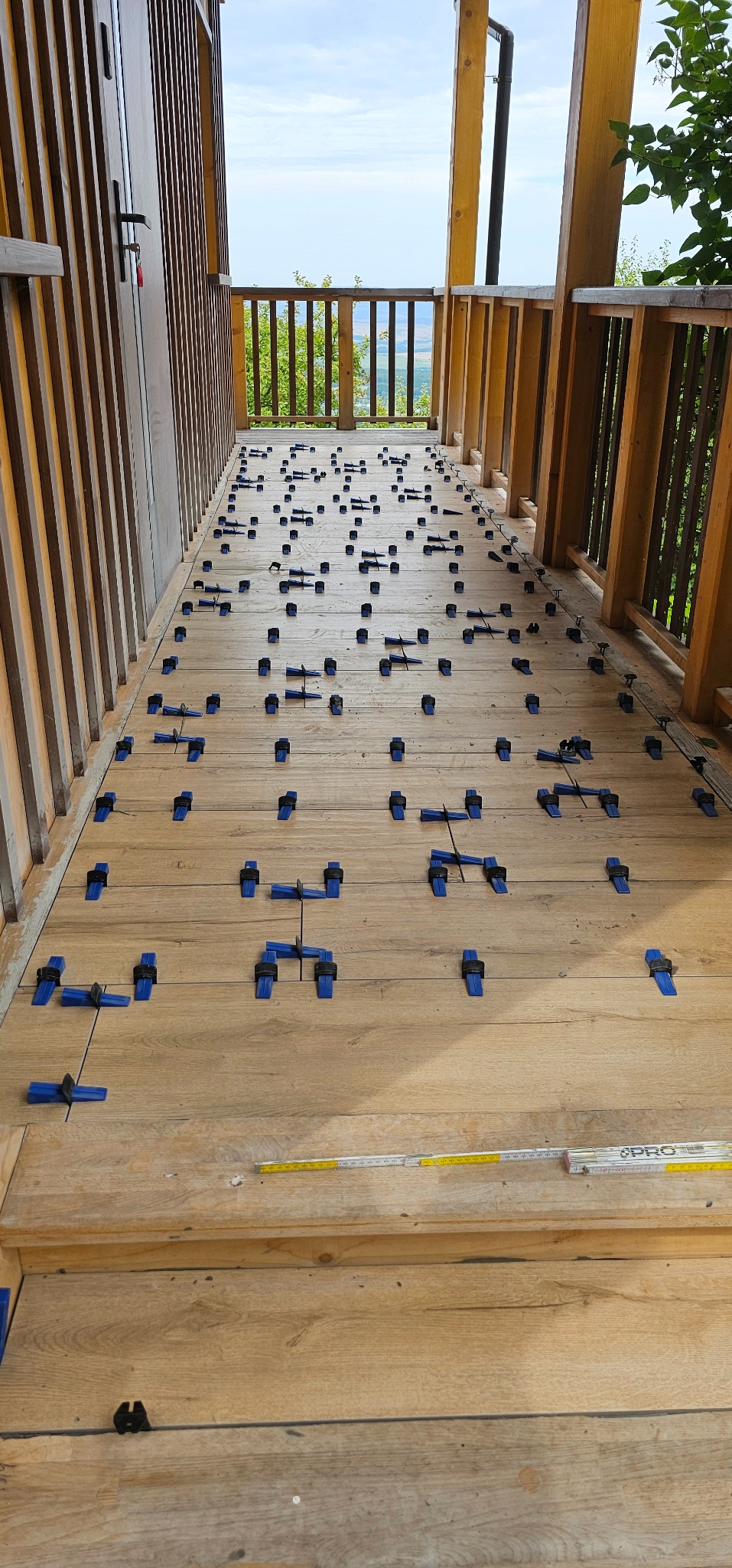 Układanie desek tarasowych na balkonie z widokiem na zieleń, z użyciem niebieskich klipsów dystansowych i miarki. Drewniana balustrada i drzwi w tle.