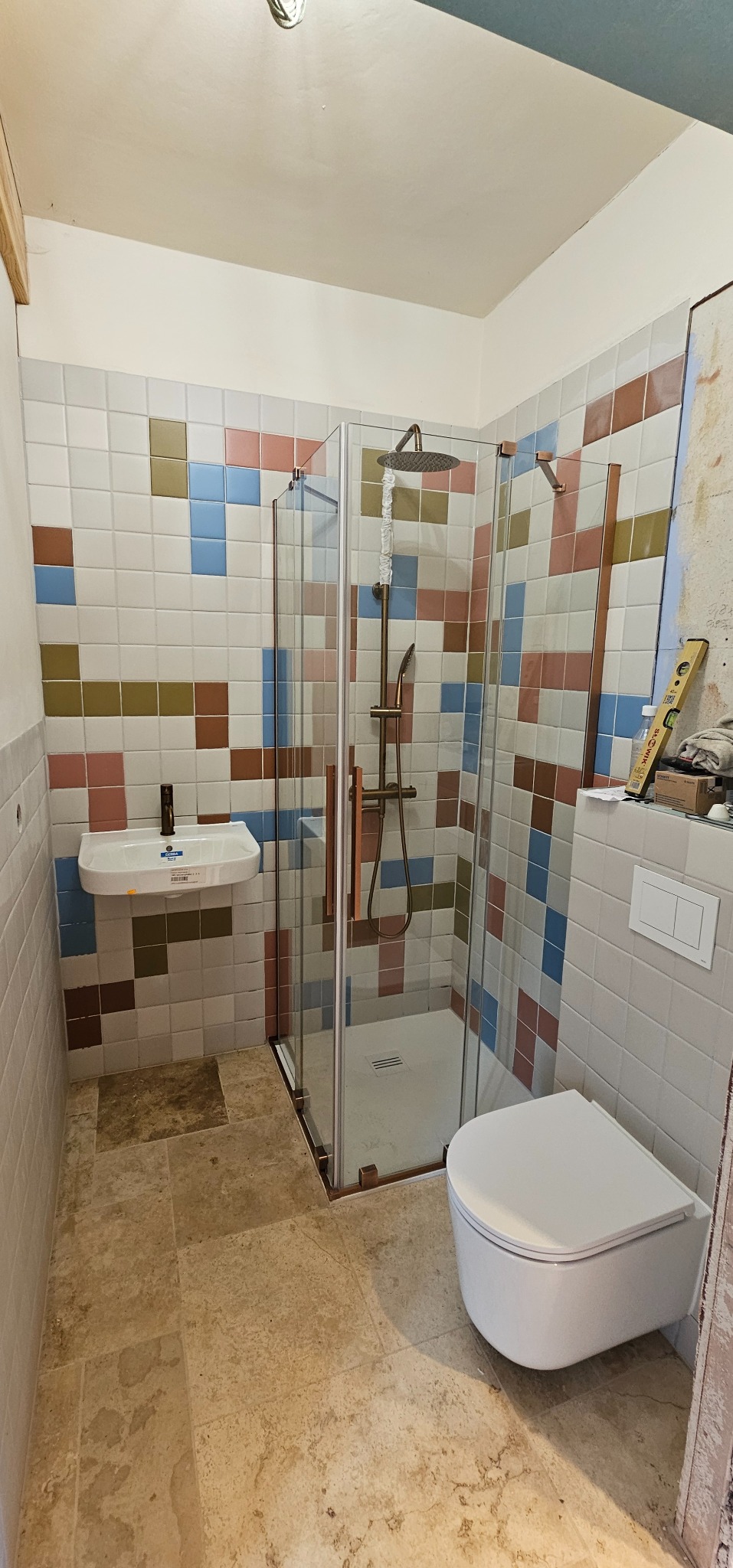 Wykończona łazienka z kabiną prysznicową, umywalką i podwieszaną toaletą. Ściany wyłożone mozaiką z kolorowych płytek, podłoga z kamiennych płyt. Elementy wykończeniowe w kolorze miedzi.