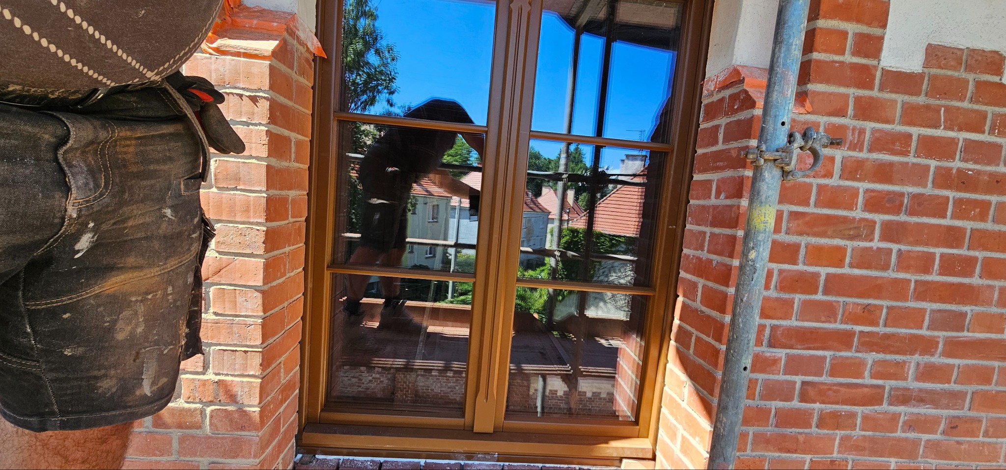 Nowe okno drewniane z szybami refleksyjnymi, zamontowane w murze z czerwonej cegły, widok z zewnątrz, fragment rusztowania, pracownik w ubraniu roboczym.