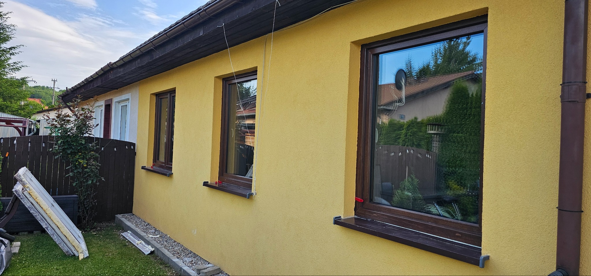 Żółta elewacja domu z trzema brązowymi oknami. W oknach odbija się zieleń drzew i niebo. Obok okien widoczna brązowa rynna. Na trawie leżą płyty ociepleniowe.