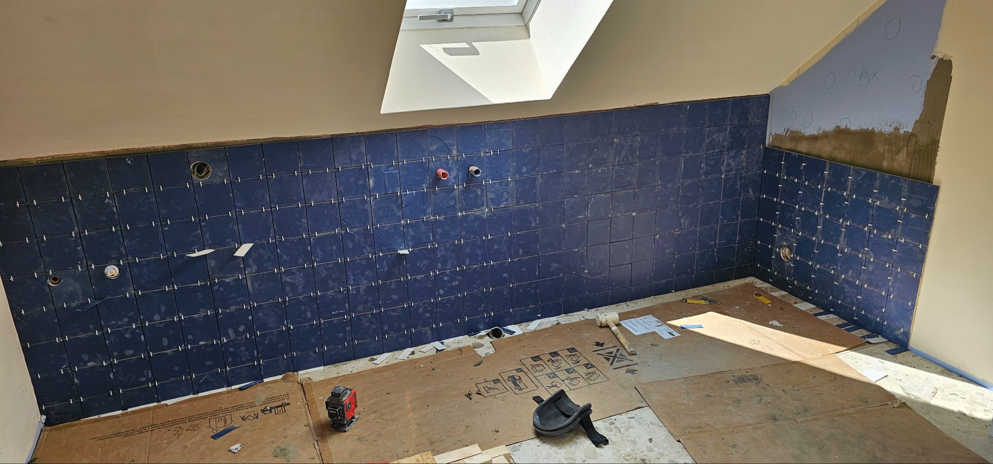 Układanie ciemnych, kwadratowych płytek w łazience na poddaszu z oknem dachowym. Widoczne krzyżyki dystansowe i narzędzia glazurnicze.